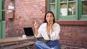 Stylish urban girl, asian woman with laptop sits in cafe, points at upper right corner banner, shows copy space with surprised face expression.