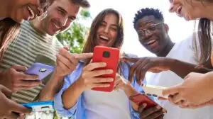 Low angle view of group smiling multiracial young students with cell watching funny reels on mobile. Gathered people laughing looking and pointing at phones. Social relations and friendship outdoors.