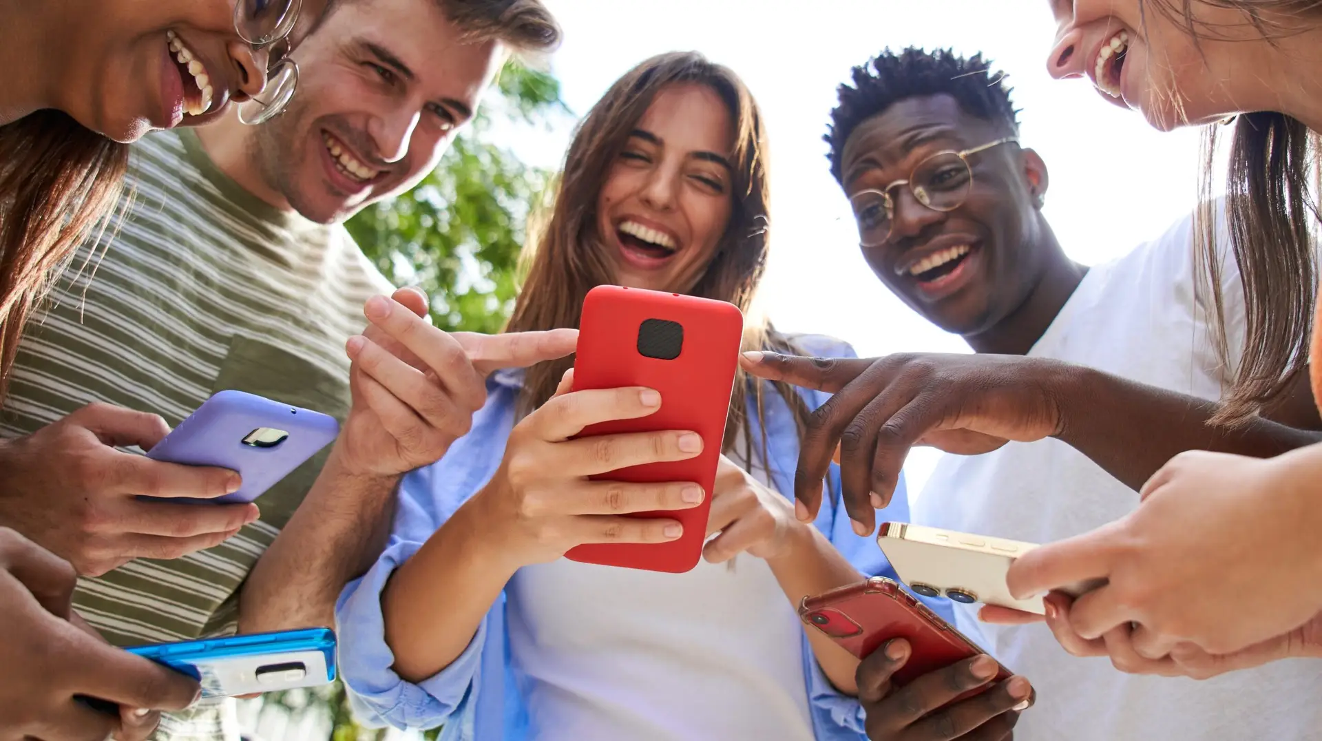 Low angle view of group smiling multiracial young students with cell watching funny reels on mobile. Gathered people laughing looking and pointing at phones. Social relations and friendship outdoors.