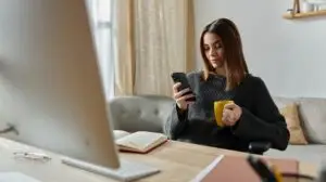 A woman works from home, glued to her phone, sipping coffee, probably managing digital marketing.