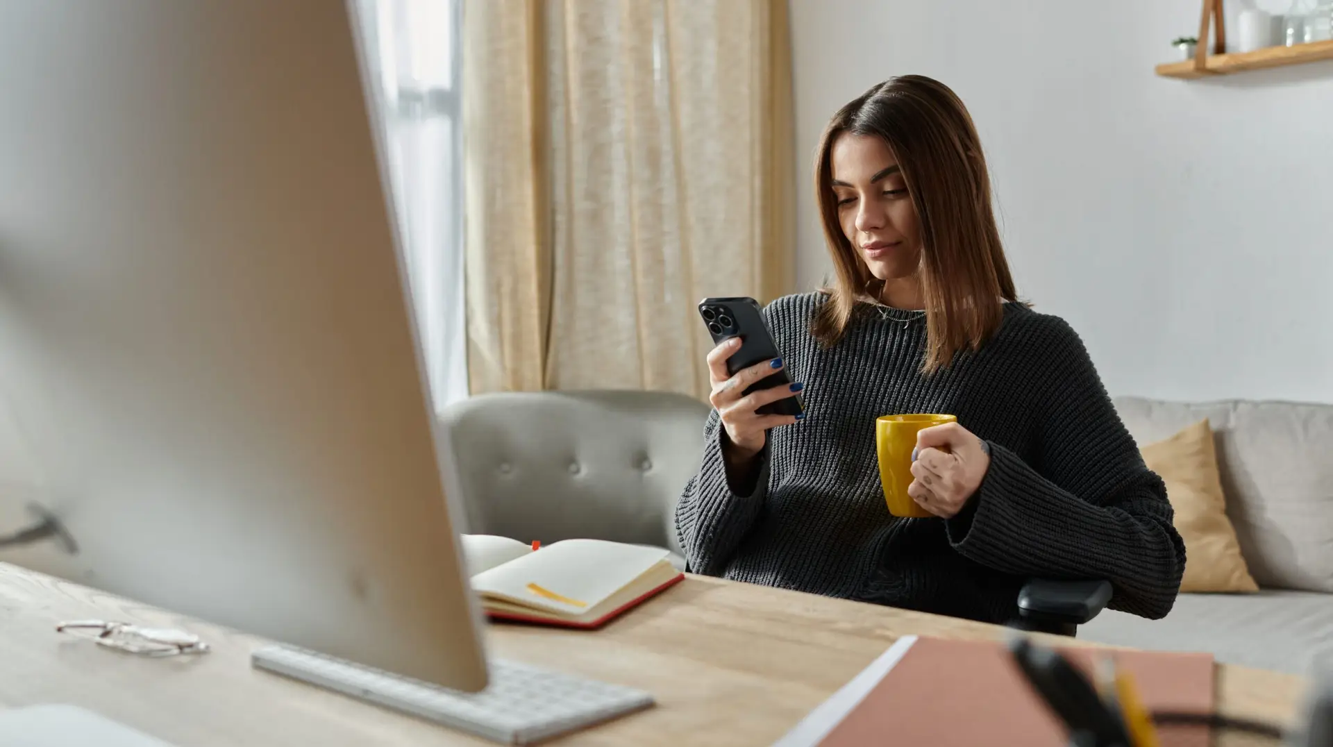 A woman works from home, glued to her phone, sipping coffee, probably managing digital marketing.